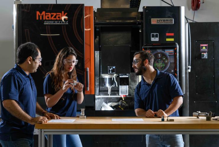 Three individuals standing at a workbench in front of a large Mazak hybrid manufacturing machine, discussing components and technical drawings.