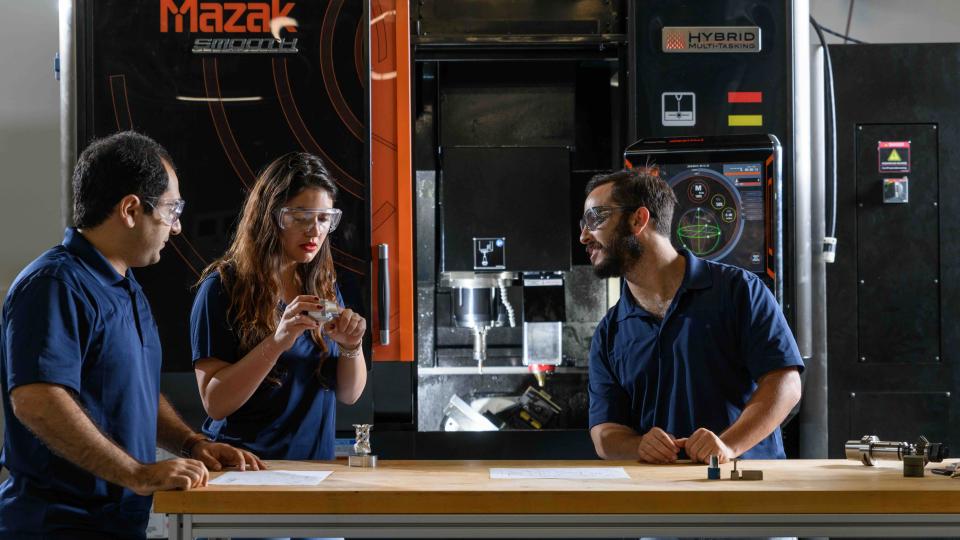 Three individuals standing at a workbench in front of a large Mazak hybrid manufacturing machine, discussing components and technical drawings.